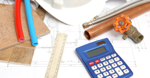 Architectural blueprint with plumbing details, alongside a white safety helmet, red and blue plastic pipes, copper fittings, a wrench, ceramic tiles, a wooden ruler, and a blue calculator for new construction plumbing cost estimation