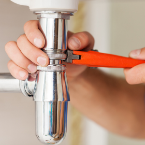 "Close-up of a plumber's hands using an orange wrench to adjust the chrome p-trap under a sink, depicting common plumbing maintenance work